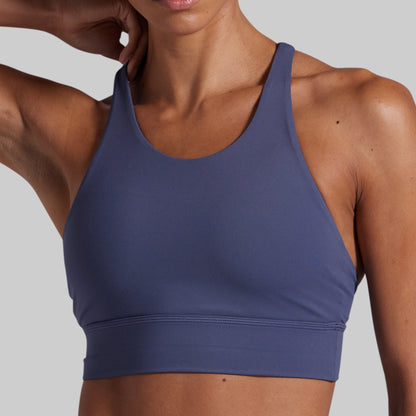 A close-up of a woman wearing a slate blue athletic sports bra against a solid gray background.