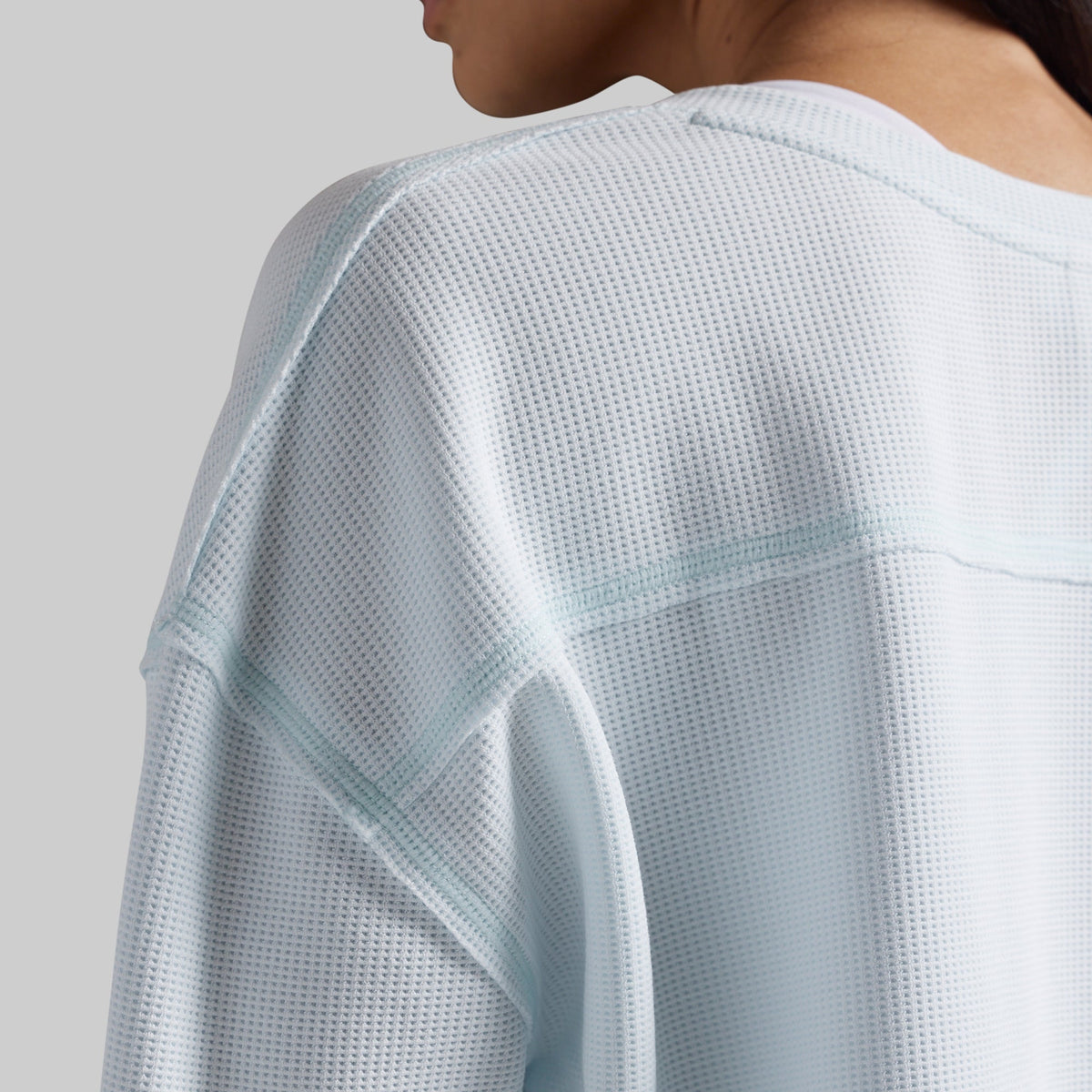 Close-up of a person wearing a light icy blue waffle-knit long-sleeve top, showing the textured fabric and shoulder seam details against a neutral gray background.