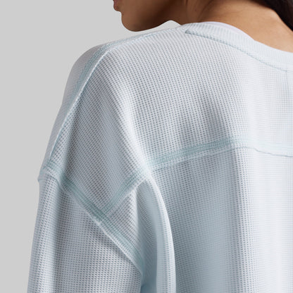 Close-up of a person wearing a light icy blue waffle-knit long-sleeve top, showing the textured fabric and shoulder seam details against a neutral gray background.
