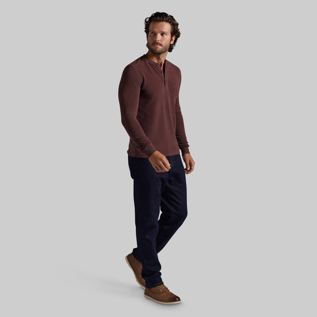 A man modeling a long-sleeve henley shirt in a deep burgundy color, paired with dark navy pants and brown suede shoes, against a solid light gray background.