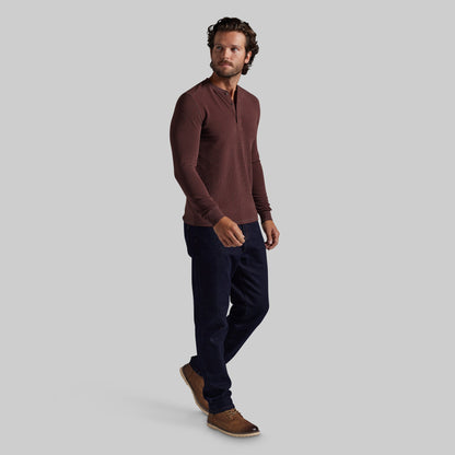 A man modeling a long-sleeve henley shirt in a deep burgundy color, paired with dark navy pants and brown suede shoes, against a solid light gray background.
