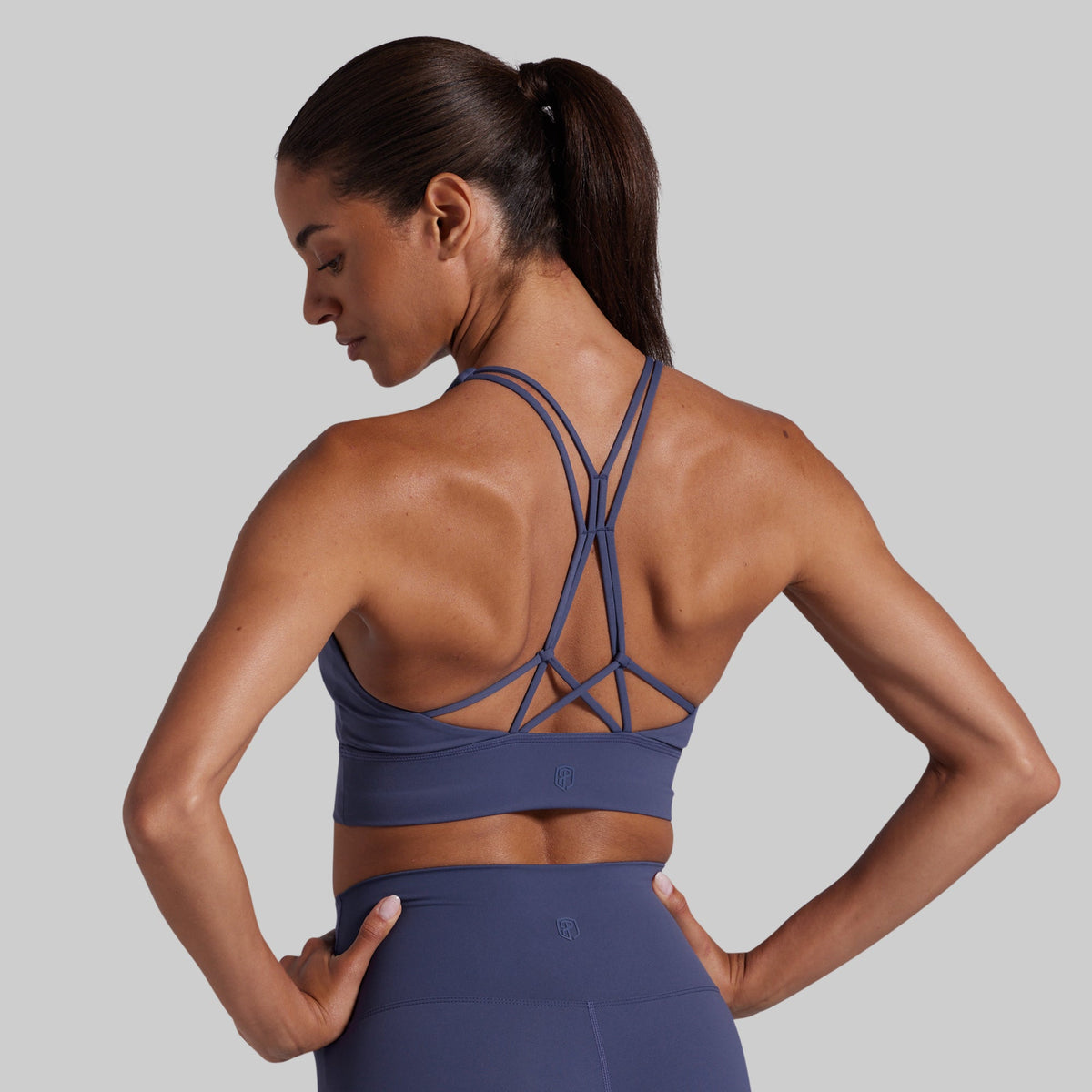 A woman shown from the back wearing a blue athletic sports bra with crisscross straps and matching high-waisted leggings against a solid gray background.