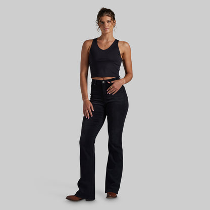 A woman modeling a black sleeveless crop top and black mid-waisted bootcut jeans against a solid light gray background.
