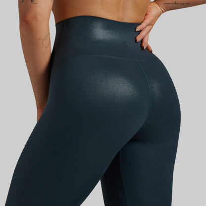 Close-up back view of a model wearing high-waisted, shiny dark teal leggings against a light gray studio background.