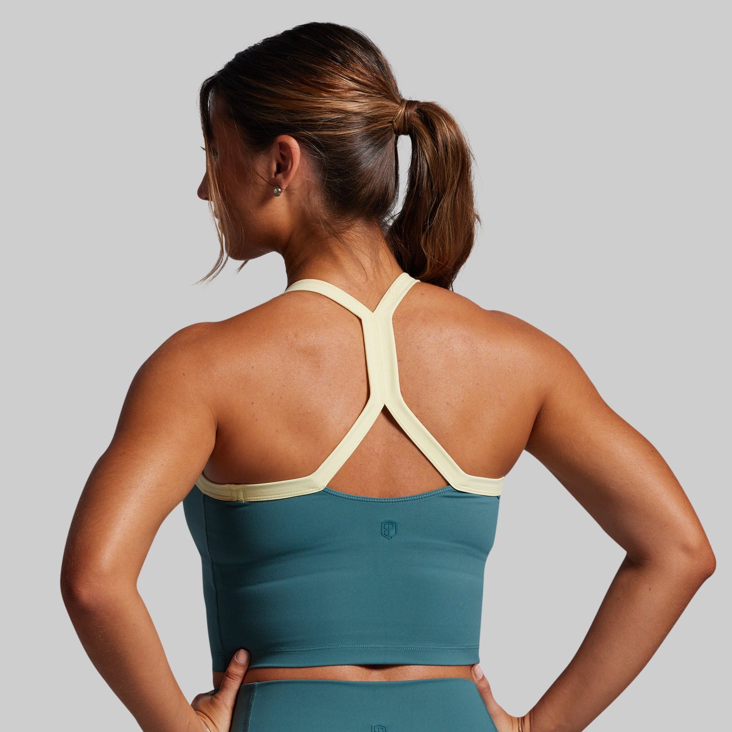 Woman wearing a teal sports bra and leggings with a yellow trim on sports bra on a gray background