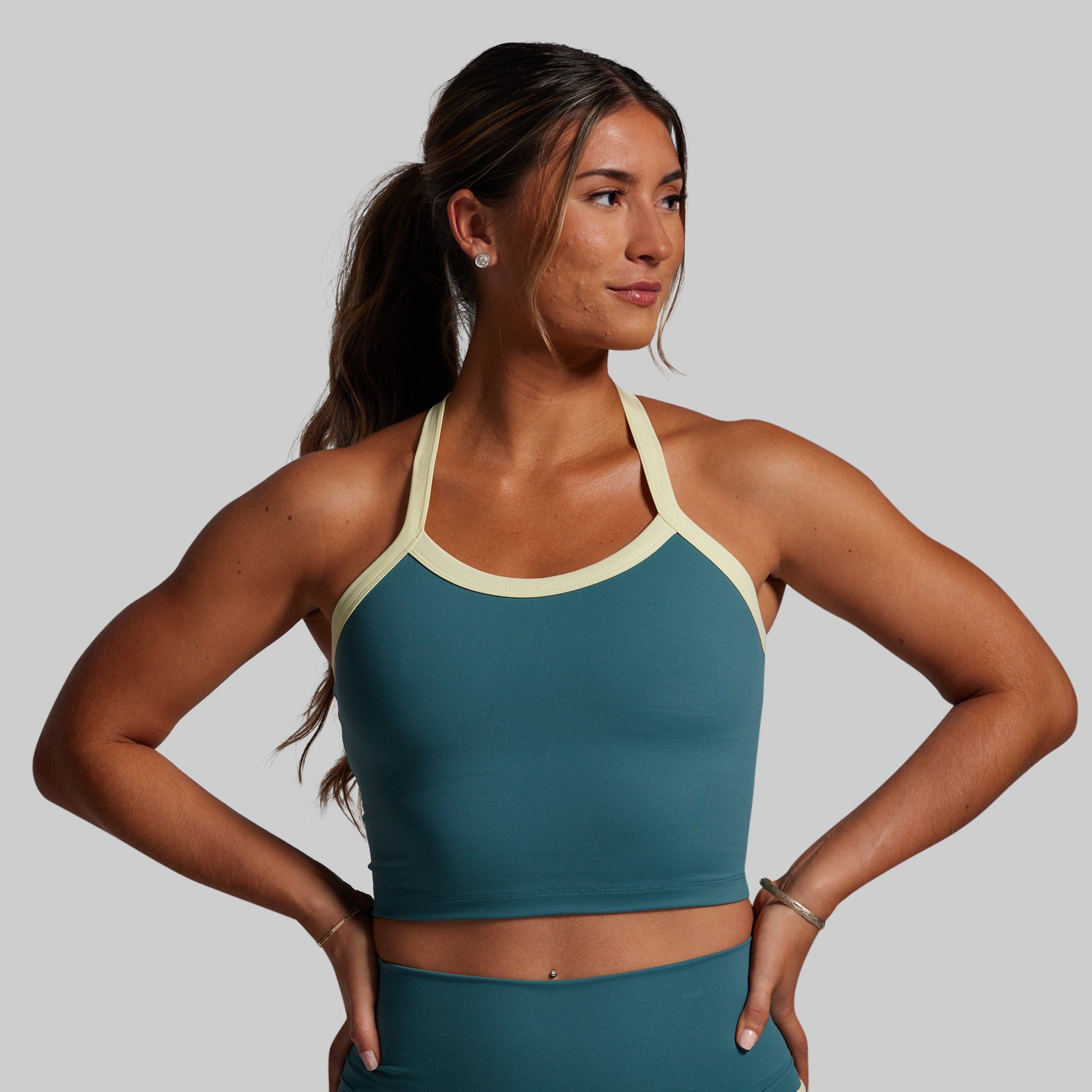 Woman wearing a teal sports bra and leggings with a yellow trim on sports bra on a gray background