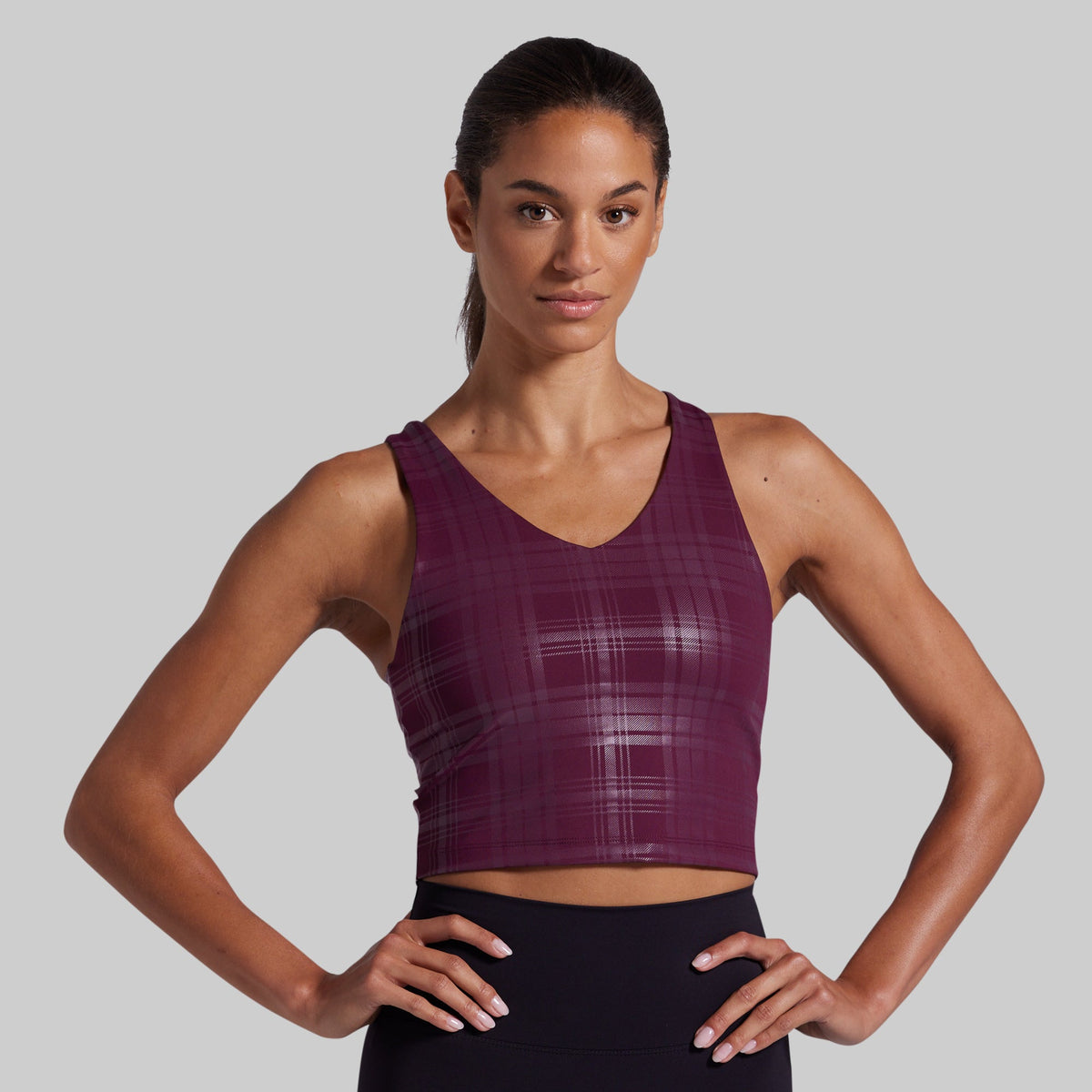 A woman modeling a sleeveless athletic sports bra in a deep plum purple color with a subtle plaid pattern, paired with black athletic bottoms, against a solid gray studio background.