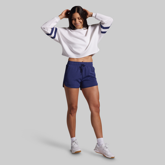 Woman wearing a white cropped sweatshirt with blue stripes and navy shorts on a gray background