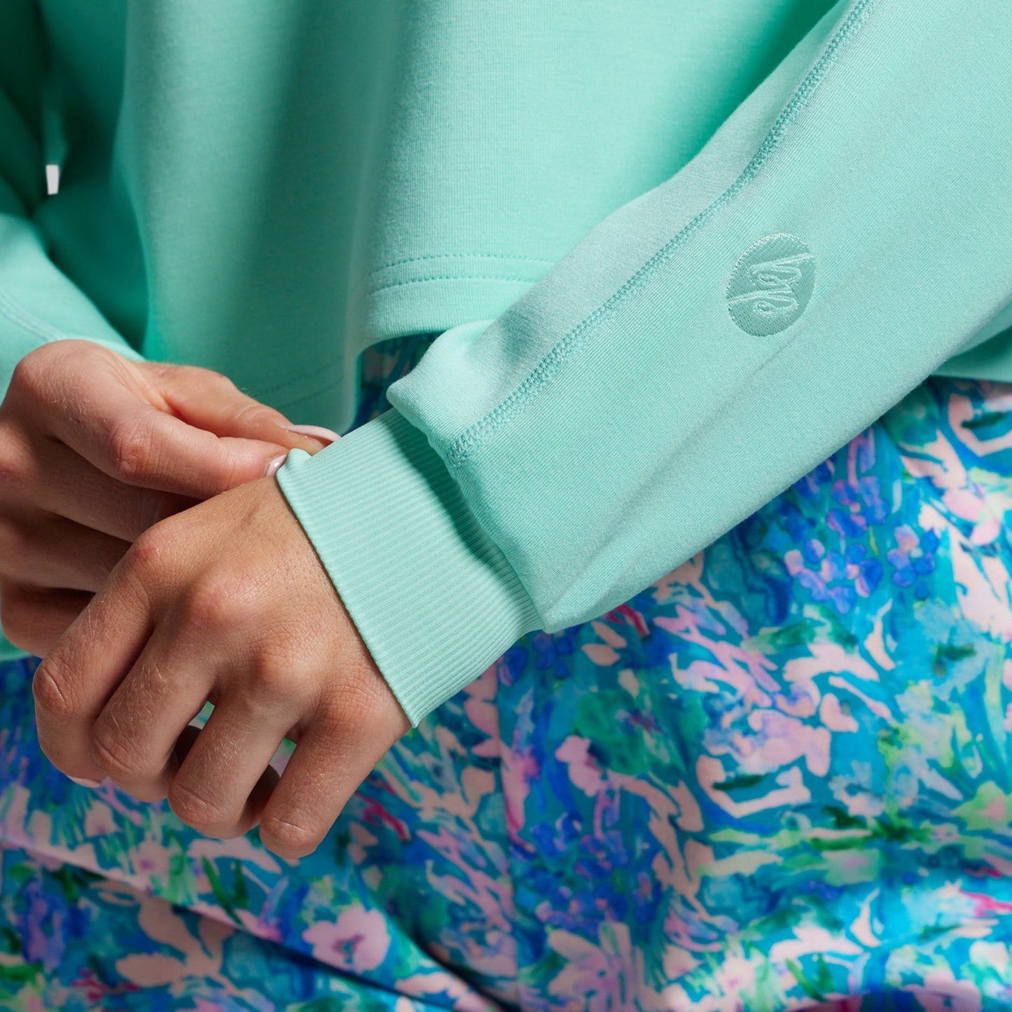 Close-up of a person wearing a light blue sweatshirt with a brand logo, against a colorful patterned background.
