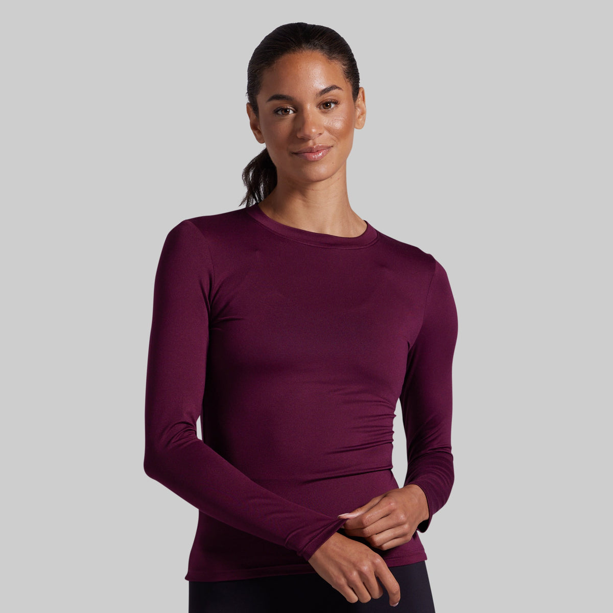 A woman modeling a long-sleeve crewneck athletic top in deep burgundy against a solid light gray background.