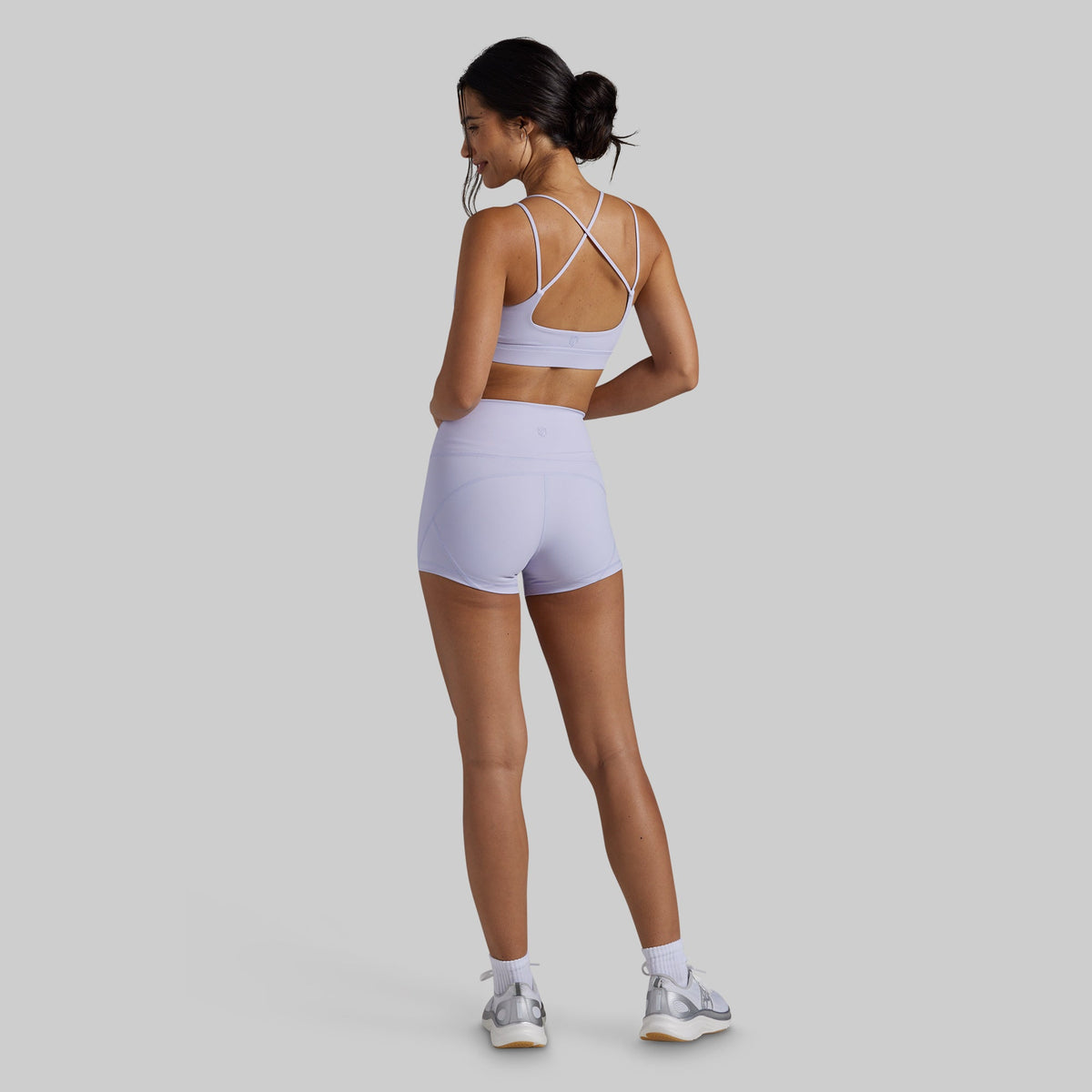 A woman wearing a light lavender athletic set consisting of a sports bra and matching high-waisted shorts, shown from the back against a solid gray background.
