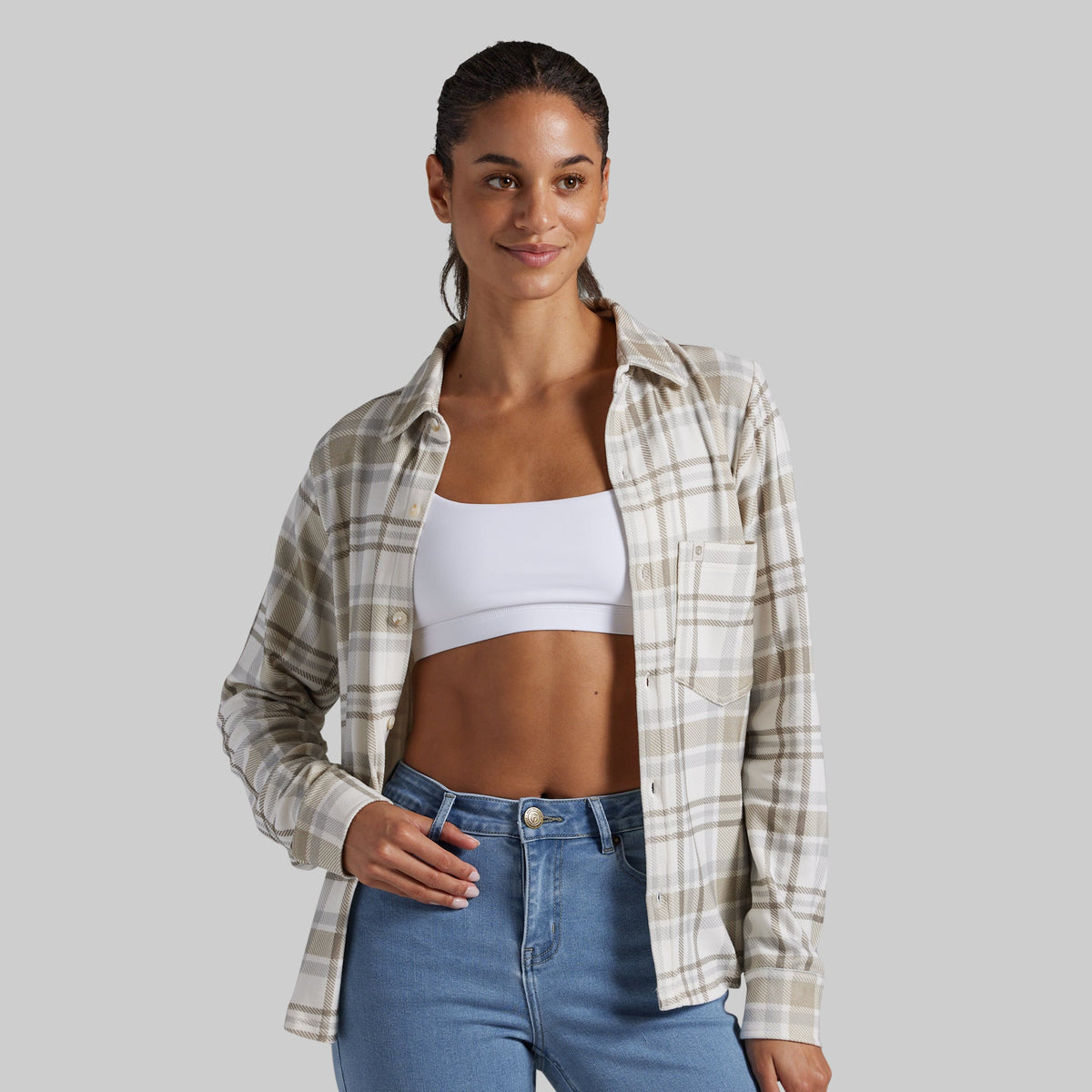 A woman modeling a long-sleeve beige and white plaid button-up shirt over a white bandeau crop top and blue jeans, set against a solid gray studio background.