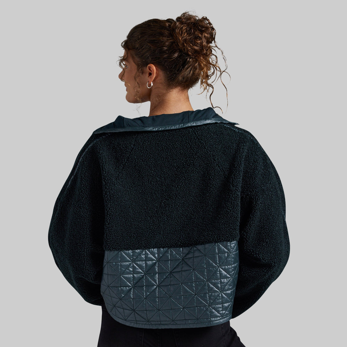 A woman shown from the back wearing a dark teal fleece jacket with a contrasting quilted geometric-patterned panel at the bottom, set against a solid gray studio background.
