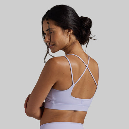 A woman shown from the back wearing a light lavender sports bra with thin crisscross straps, against a solid light gray background.