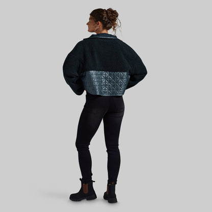 A woman is shown from the back wearing a dark teal cropped jacket with a textured geometric quilted panel across the lower back, paired with black leggings and black ankle boots, against a solid gray background.