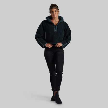 A woman modeling a dark teal fleece pullover with a half-zip neckline and ribbed cuffs, paired with black pants and black ankle boots, against a solid light gray background.