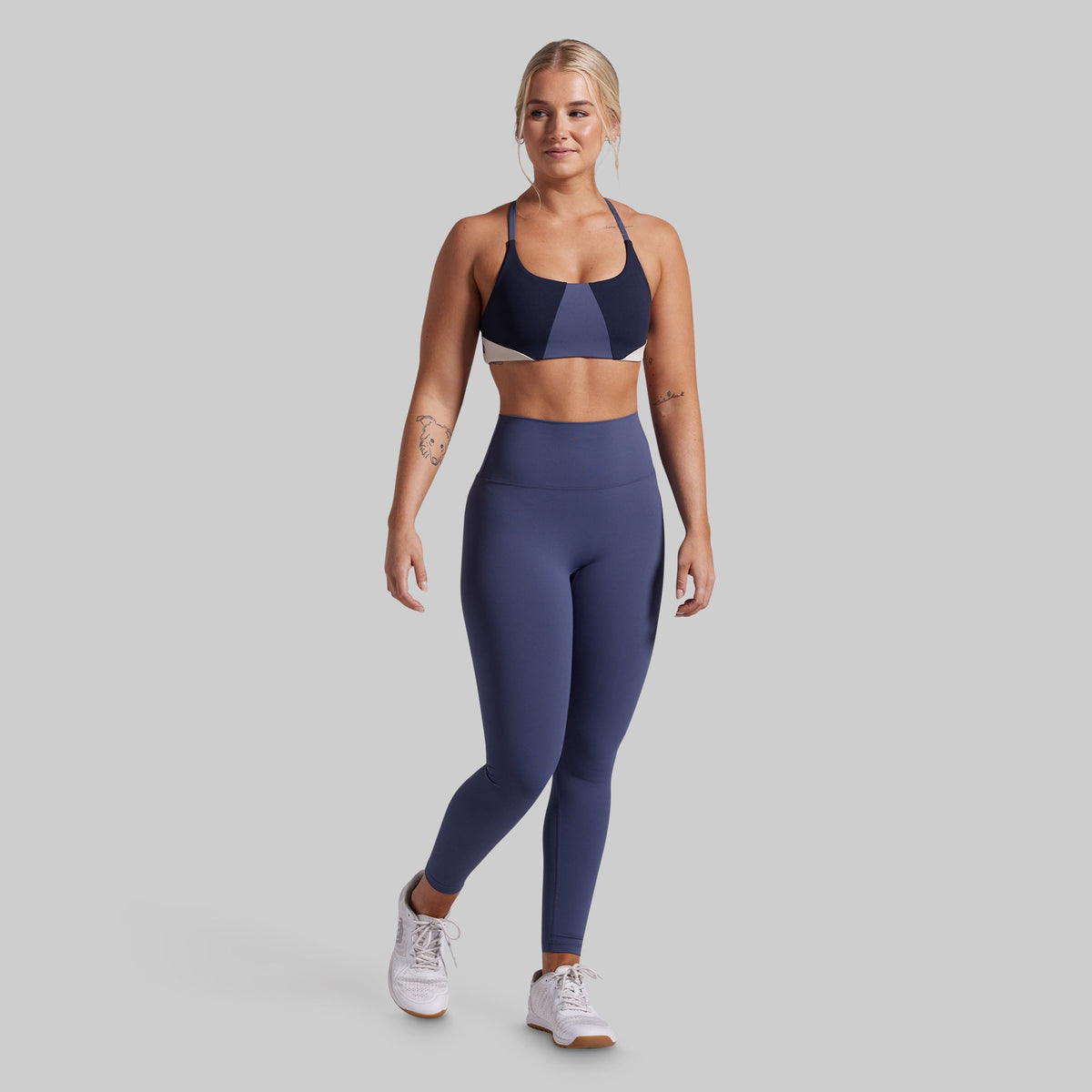 A woman modeling a matching athletic set consisting of a color-blocked sports bra and high-waisted leggings in navy, blue, and light gray, paired with white sneakers, against a solid gray background.