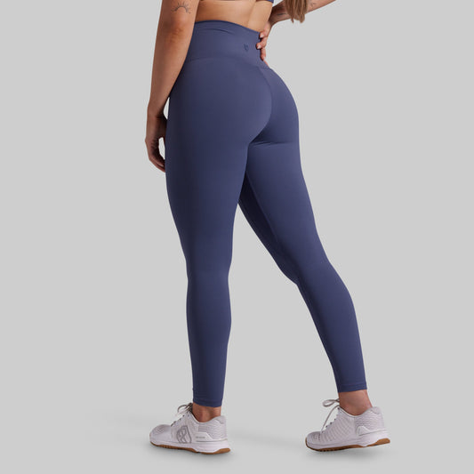 A woman wearing high-waisted, full-length athletic leggings in a muted slate blue color, shown from a rear three-quarter angle against a solid light gray background.