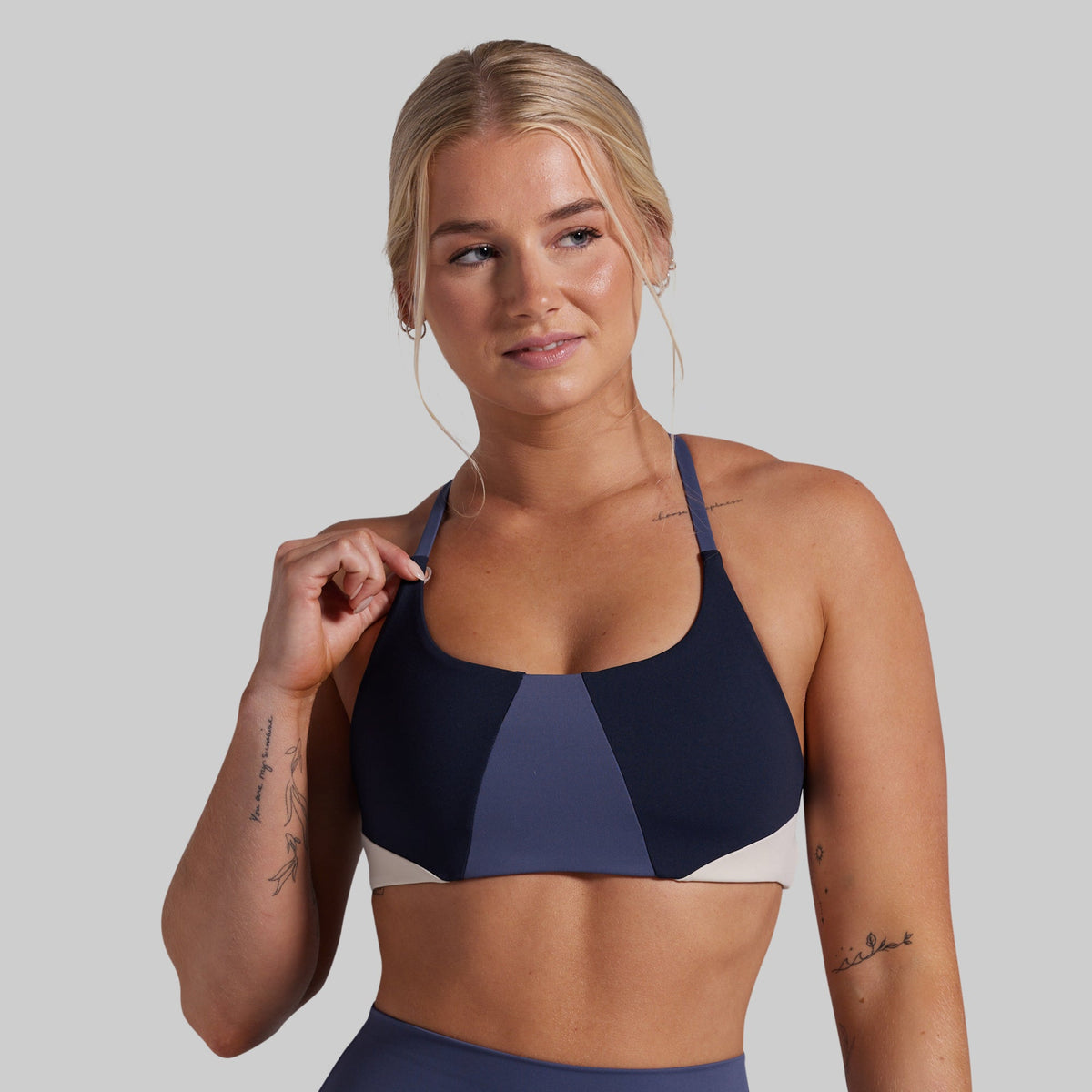 A blonde model wearing a navy blue colorblock sports bra with a central navy panel and a light beige underband, shown against a solid gray background.