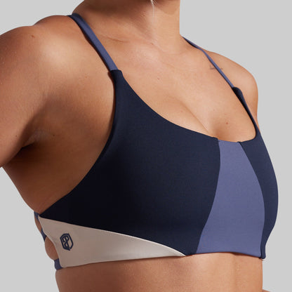 Close-up of a woman wearing a color-blocked athletic sports bra in navy, slate blue, and white, with a small geometric logo on the white side band.