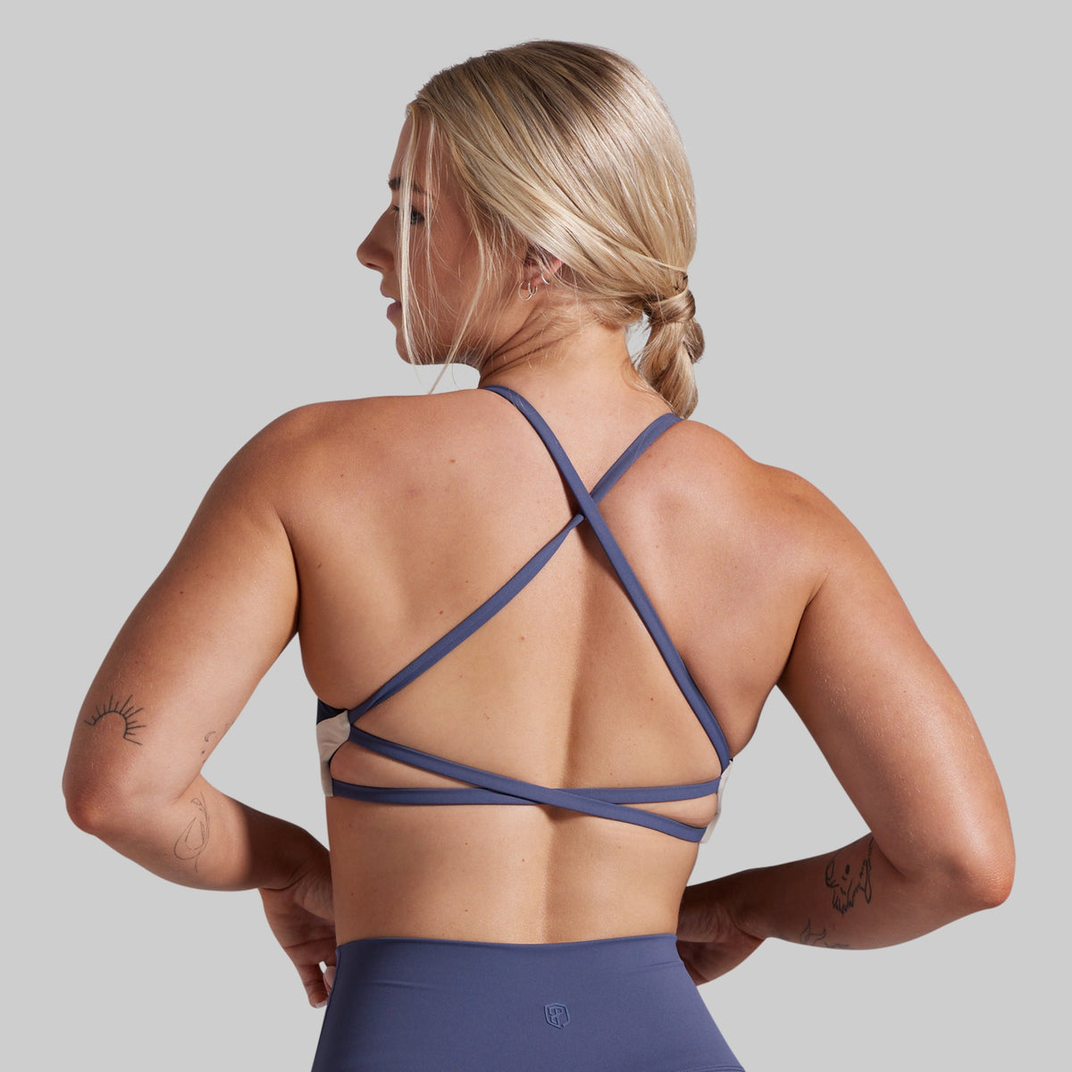 A woman shown from the back wearing a navy blue athletic sports bra with crisscross straps, paired with matching high-waisted bottoms, against a solid gray background.