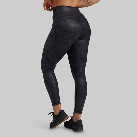 Rear three-quarter view of a model wearing high-waisted, full-length athletic leggings with a dark charcoal gray and black mottled texture, paired with black sneakers, against a solid light gray background.