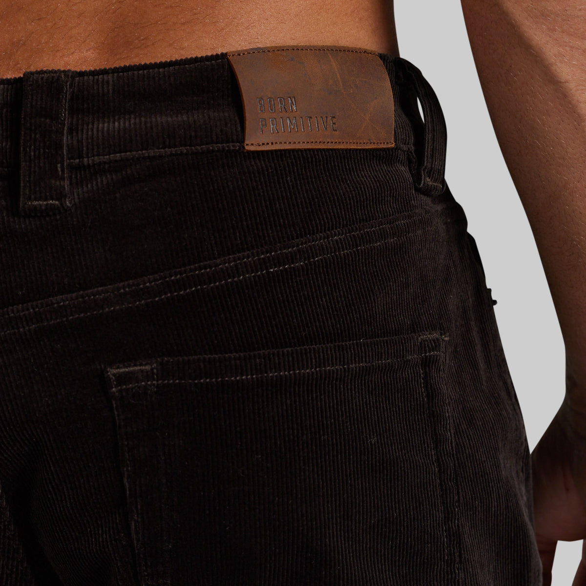 Close-up of the back waistband of dark brown corduroy pants worn by a model, showing a brown leather brand patch that reads 'BORN PRIMITIVE'.