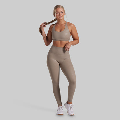 A woman modeling a matching athletic set consisting of a sports bra and high-waisted leggings in a taupe color, paired with white sneakers, against a solid light gray background.