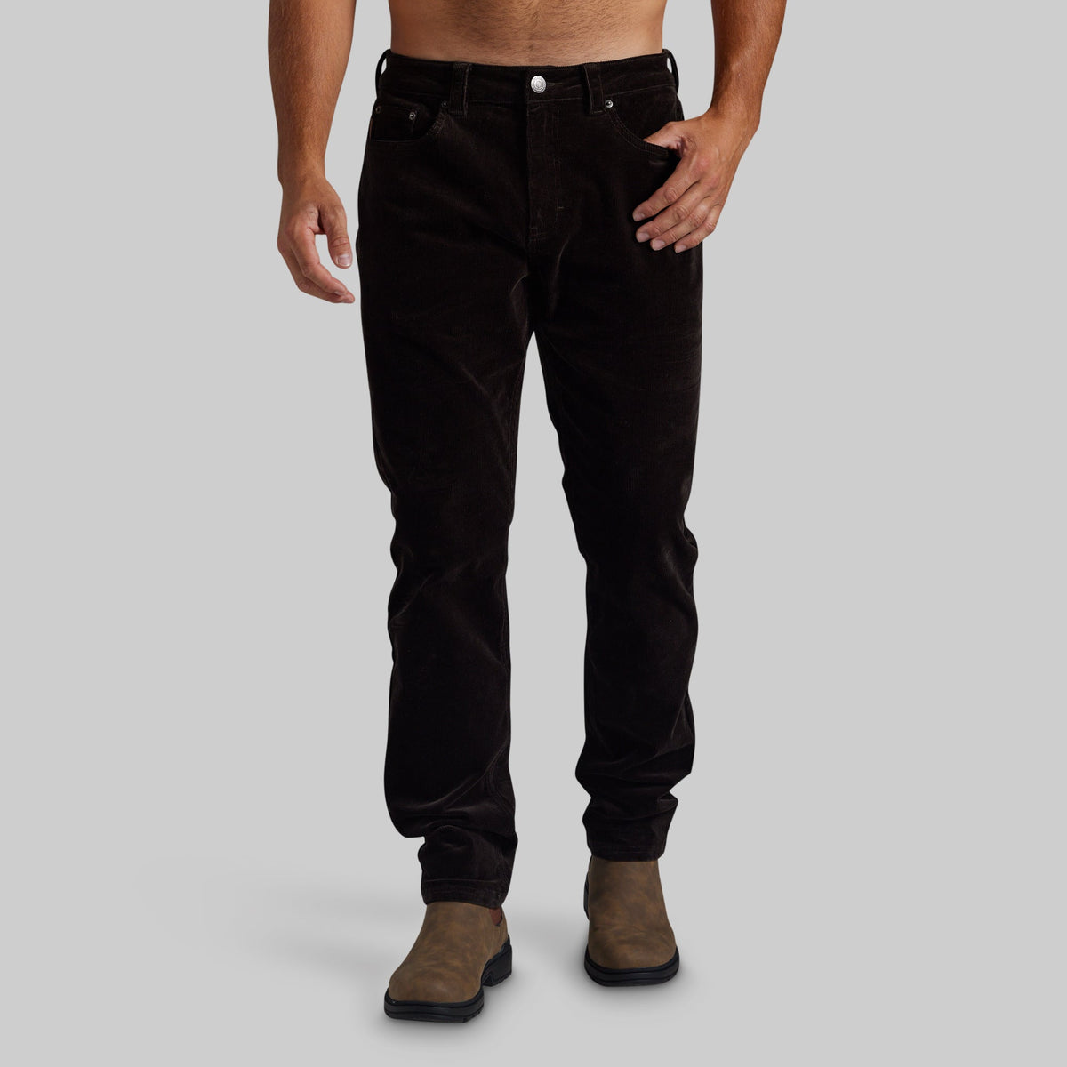 A person modeling slim-fit dark brown corduroy pants with brown suede boots, shown from the waist down against a plain light gray studio background.