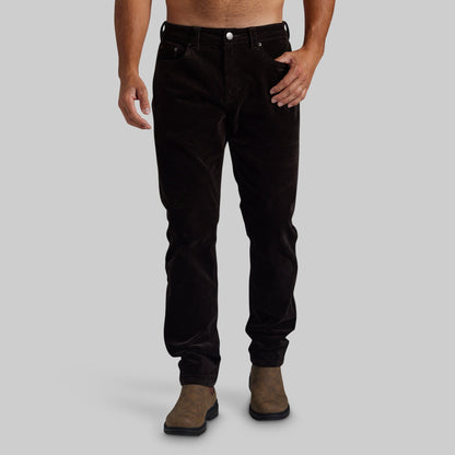 A person modeling slim-fit dark brown corduroy pants with brown suede boots, shown from the waist down against a plain light gray studio background.