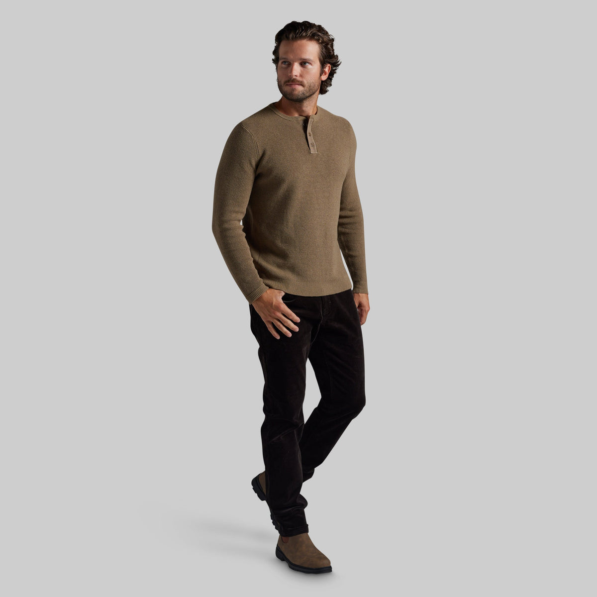 A man modeling a long-sleeve henley sweater in a warm taupe-brown color, paired with black pants and brown boots, against a solid light gray background.