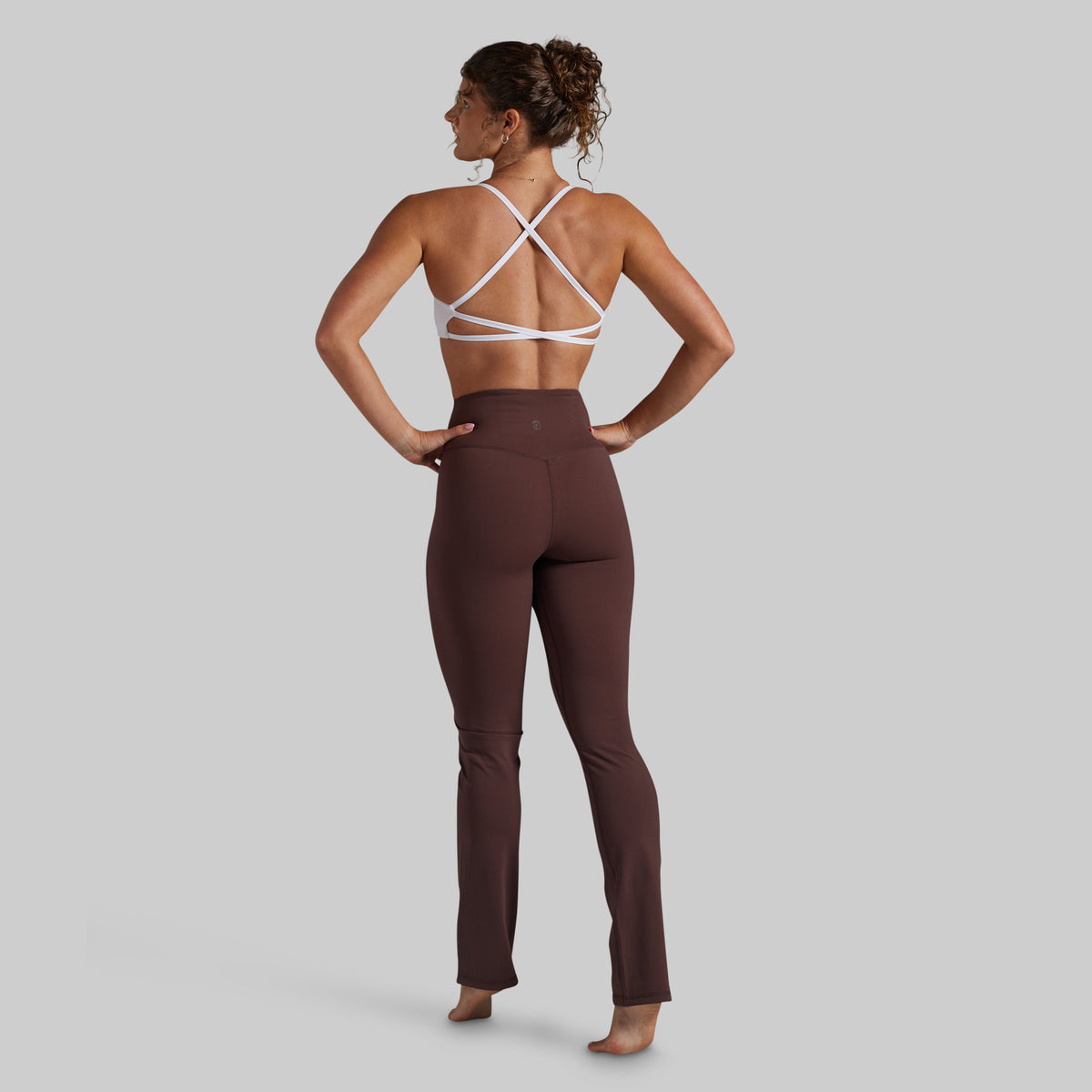 A woman shown from the back wearing high-waisted brown yoga pants and a white strappy sports bra, standing against a solid light gray background.
