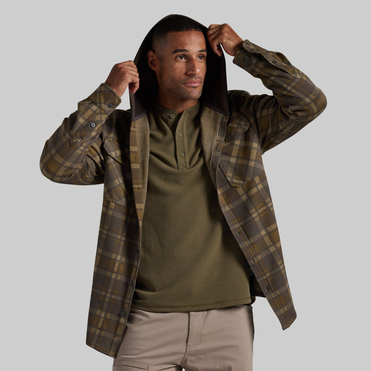 A man wearing a brown and olive green plaid hooded flannel shirt over an olive green henley shirt and khaki pants, posing with the hood up against a solid gray background.