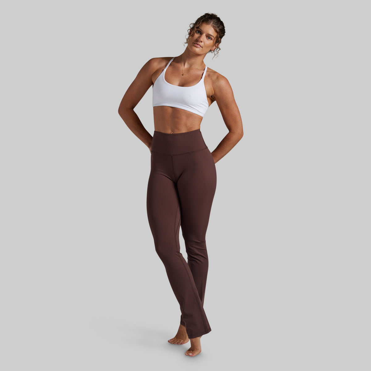 A woman modeling high-waisted, ankle-length athletic yoga pants in a dark brown color, paired with a white sports bra, against a solid light gray background.
