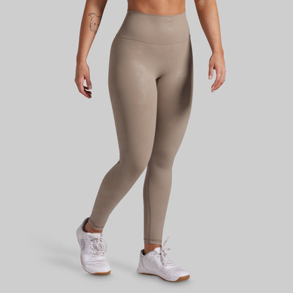 A model wearing high-waisted, full-length athletic leggings in a taupe-beige color, paired with white sneakers, shown from the waist down against a solid light gray background.
