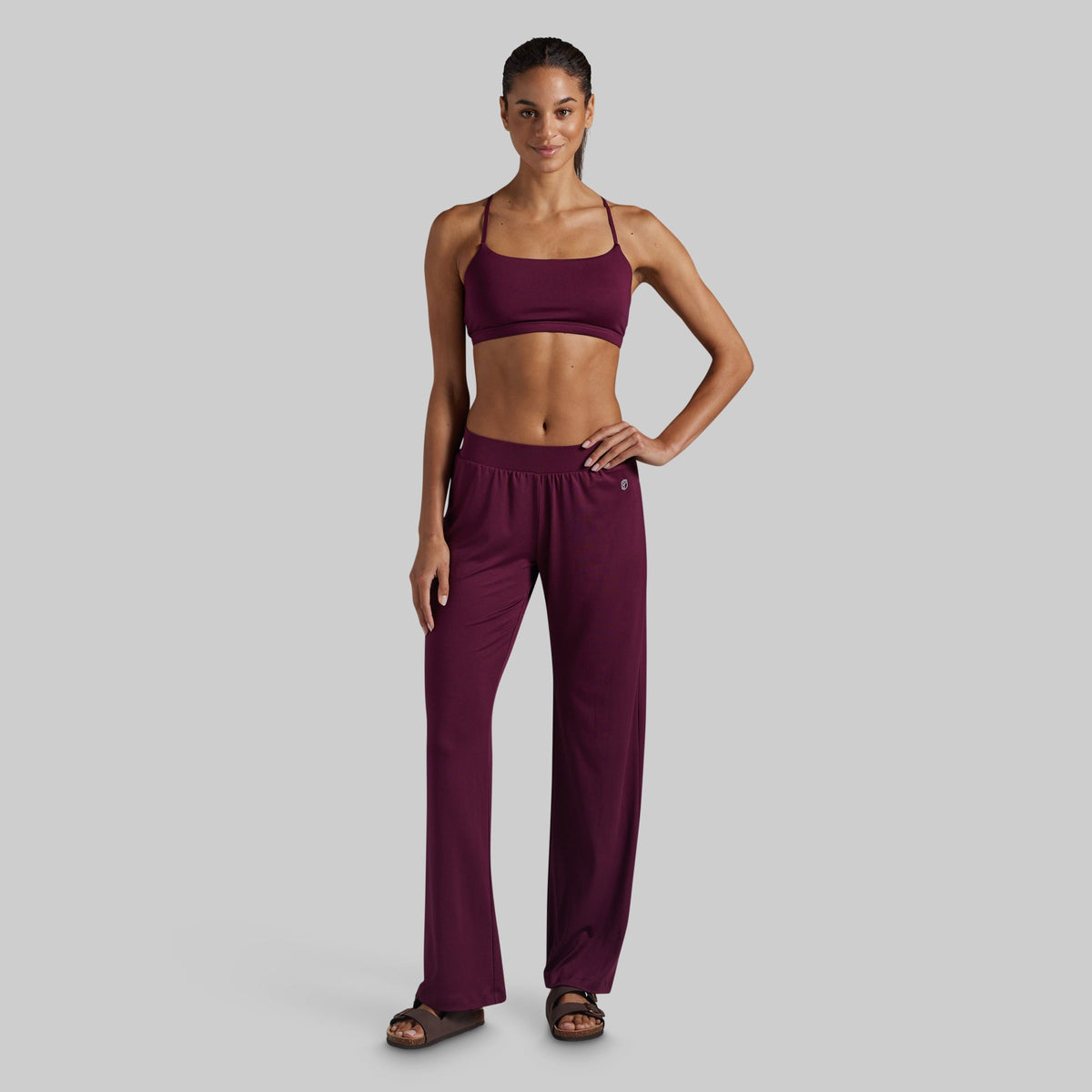 A woman modeling a matching two-piece loungewear set in deep burgundy, featuring a spaghetti-strap lounge bra and high-waisted wide-leg pants, set against a solid light gray background.