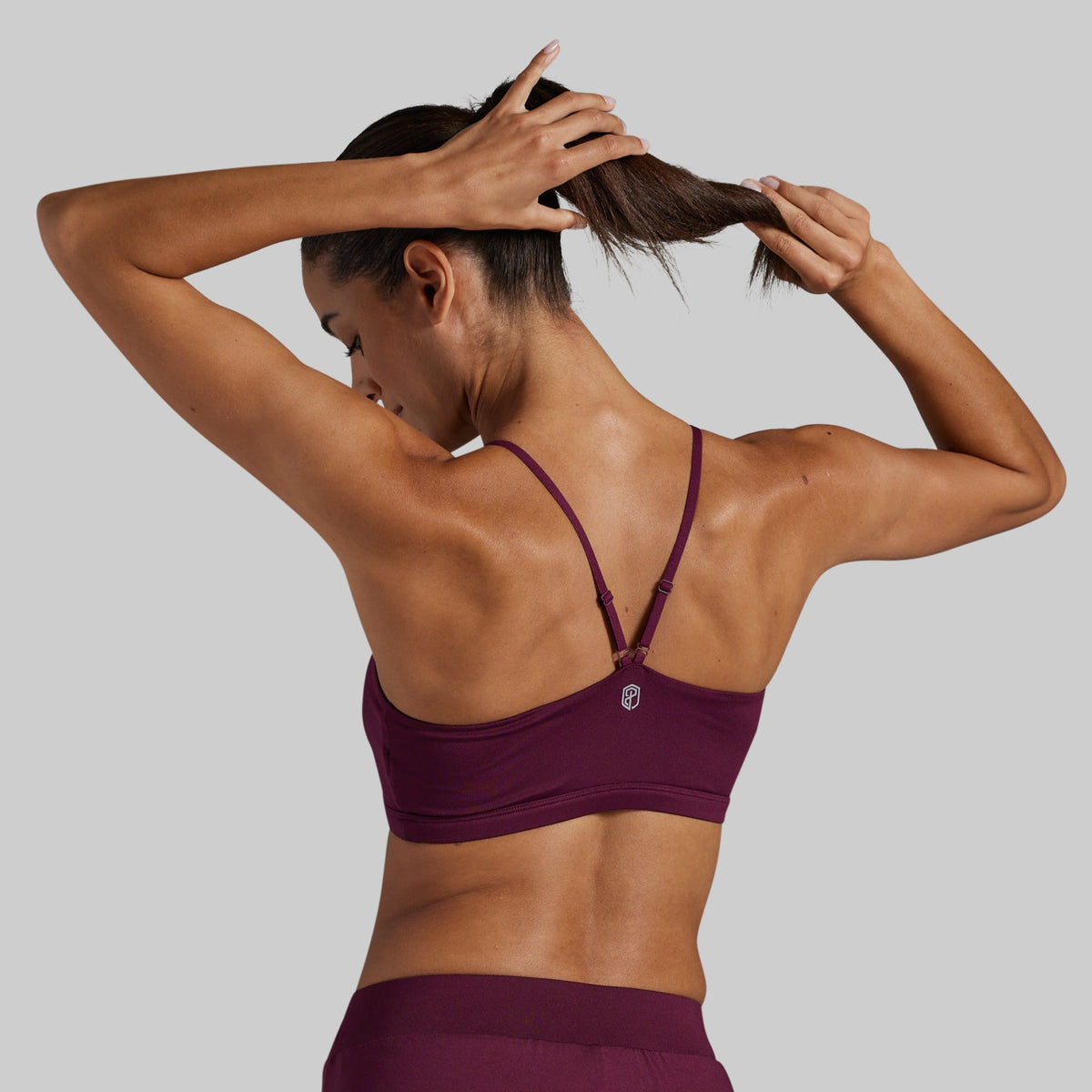 A woman shown from the back wearing a deep plum-purple lounge bra, with her hands adjusting her hair. 