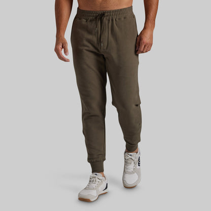 A man wearing olive green jogger sweatpants with a drawstring waist and elastic cuffs, paired with white sneakers, shown from the waist down against a light gray studio background.