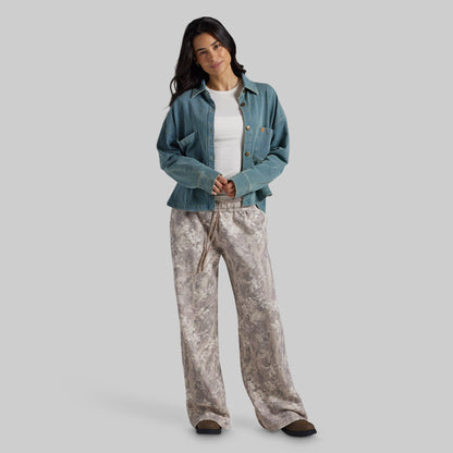 A woman modeling a casual outfit consisting of a teal cropped denim jacket over a white top and wide-leg beige and gray patterned pants, set against a solid gray studio background.