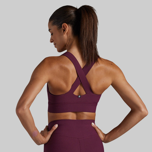 A woman shown from the back wearing a ribbed athletic sports bra and matching high-waisted leggings in a deep plum purple color, set against a solid light gray background.