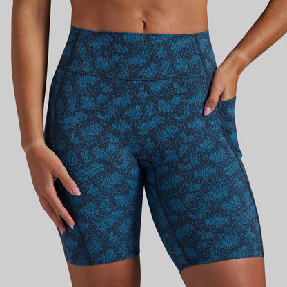 A close-up of a model wearing high-waisted athletic biker shorts with a blue and black abstract pattern, shown from the waist to mid-thigh against a solid gray background.