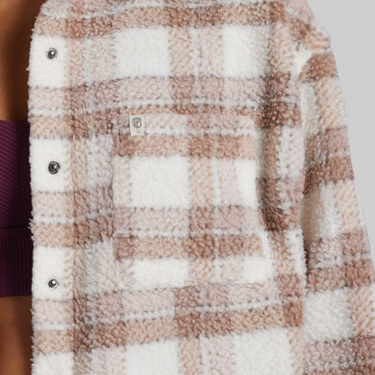 Close-up of a sherpa fleece button-up shirt jacket with a brown and white plaid pattern, featuring a chest pocket and snap buttons.