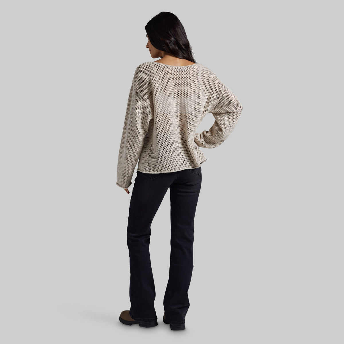 A woman shown from the back wearing a cream-colored, textured knit long-sleeve sweater and black pants, standing against a solid light gray background.