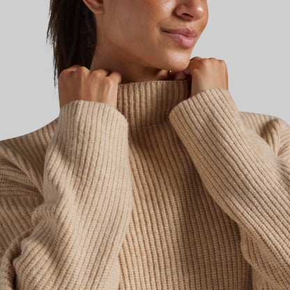 Close-up of a person wearing a chunky ribbed turtleneck sweater in a warm beige color, adjusting the collar with both hands against a plain light gray background.