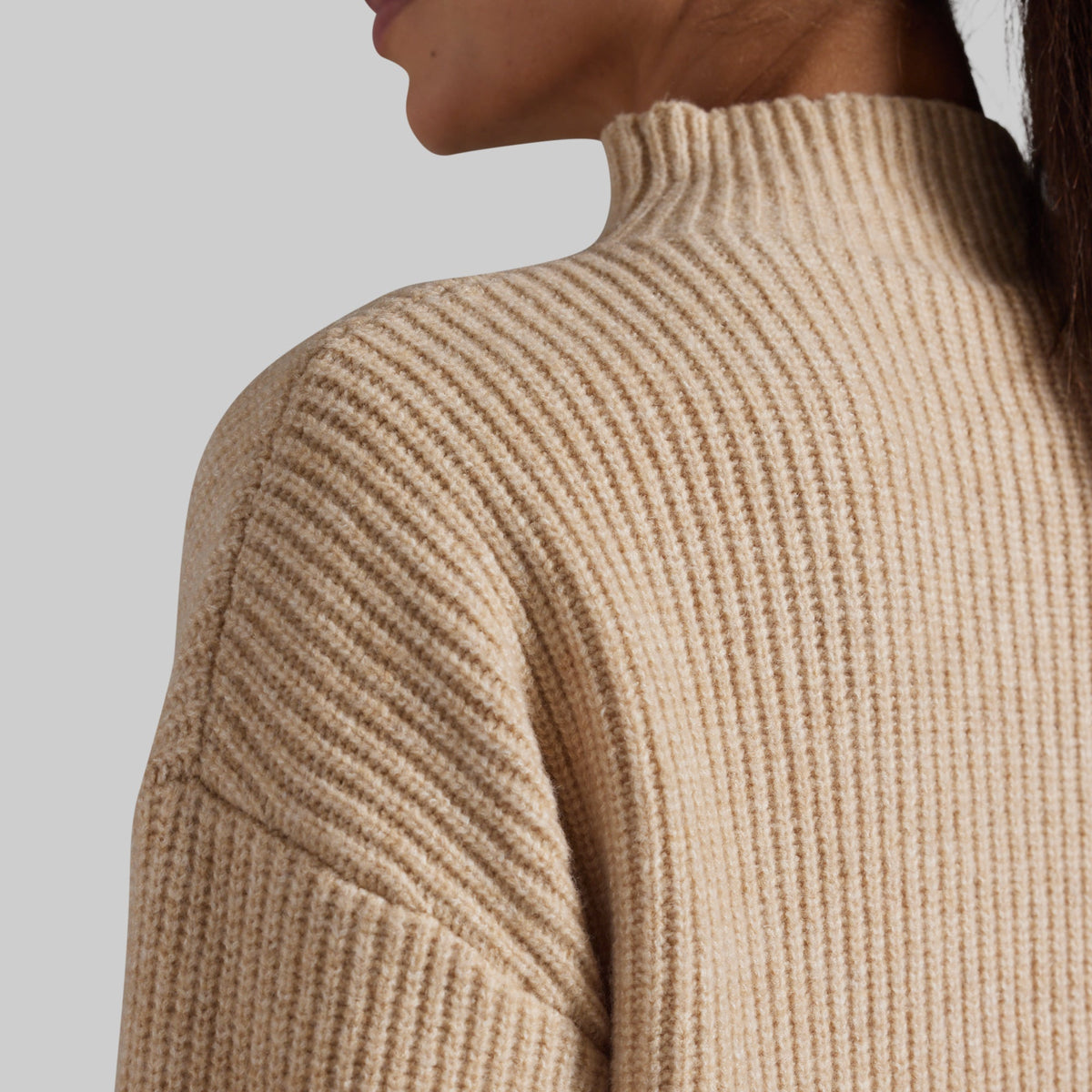 Close-up of a person wearing a ribbed knit mock neck sweater in a warm beige color, showing the texture and shoulder detail against a light gray background.