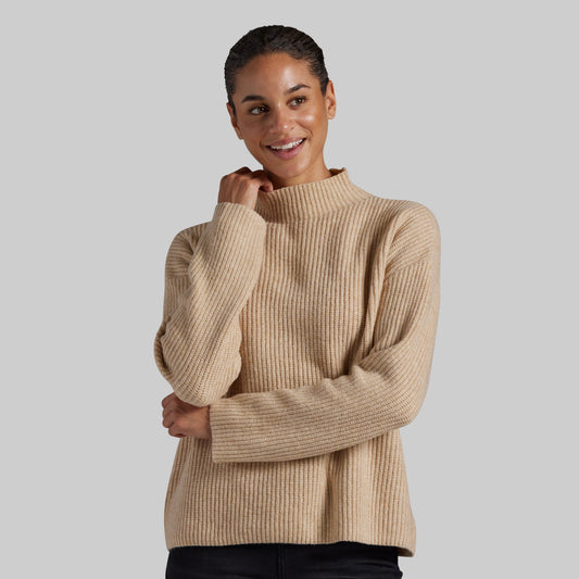 A woman wearing a beige ribbed knit mock neck sweater, posing against a solid gray studio background.