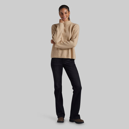 A woman modeling a beige ribbed knit mock-neck sweater and black jeans against a solid light gray background.