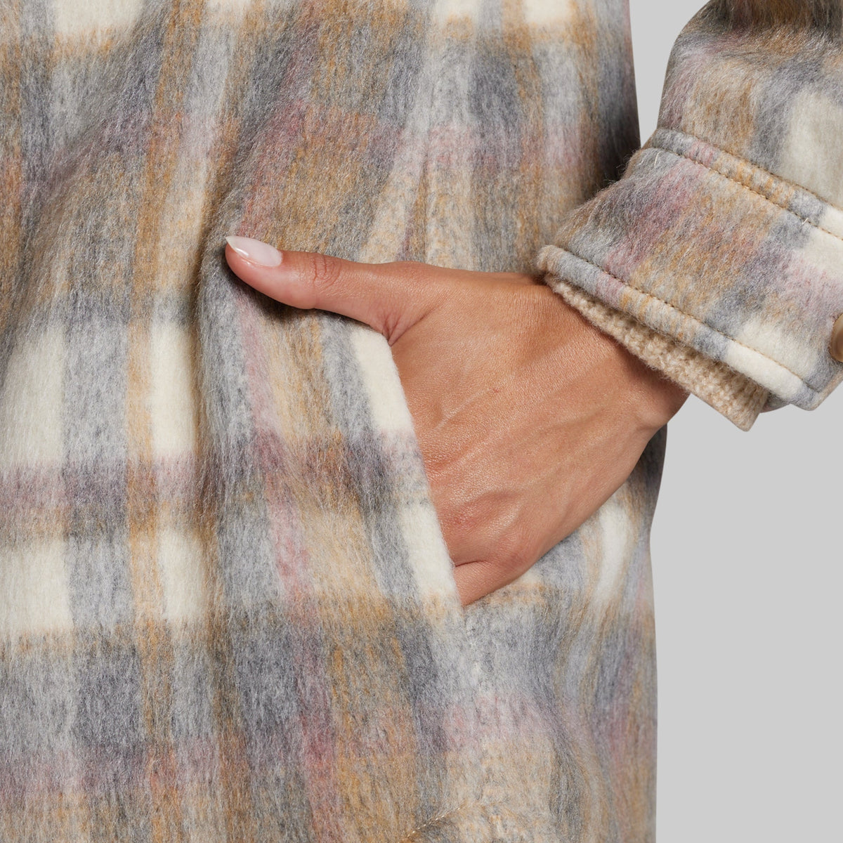 Close-up of a person wearing a fuzzy plaid coat with a hand in the pocket, showing the sleeve cuff and textured fabric against a light gray background.