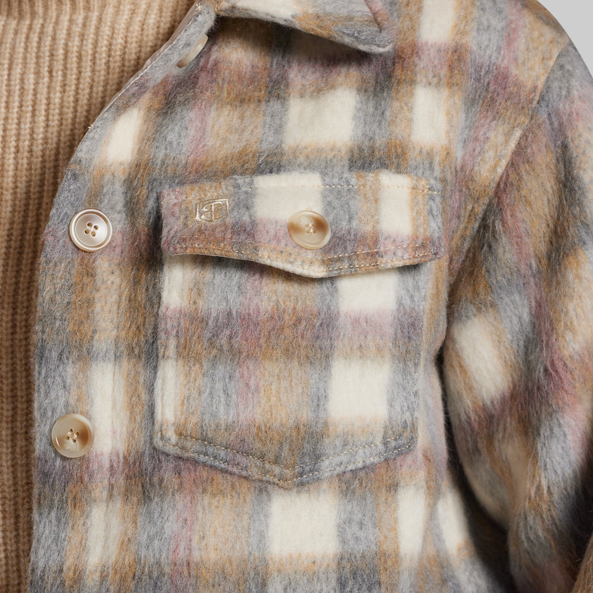 Close-up of a fuzzy plaid jacket with a chest pocket, featuring a cream, gray, and muted pink pattern, worn over a tan ribbed knit sweater.