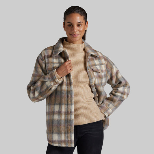A woman modeling a beige and brown plaid button-up flannel jacket over a ribbed cream turtleneck sweater and black pants, set against a solid gray studio background.